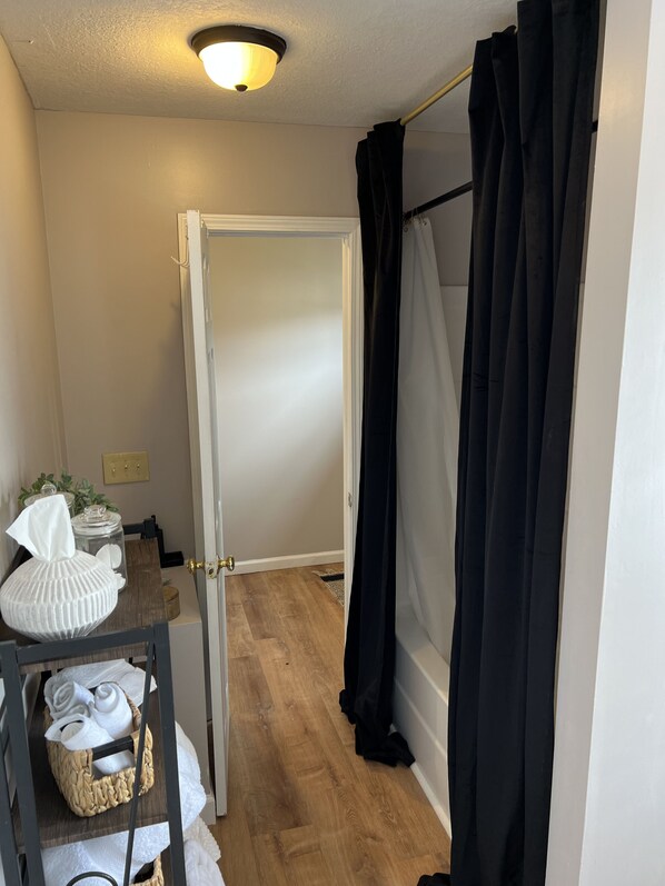 A bathroom with a wooden floor, a white door, a black shower curtain, and a towel rack with towels.