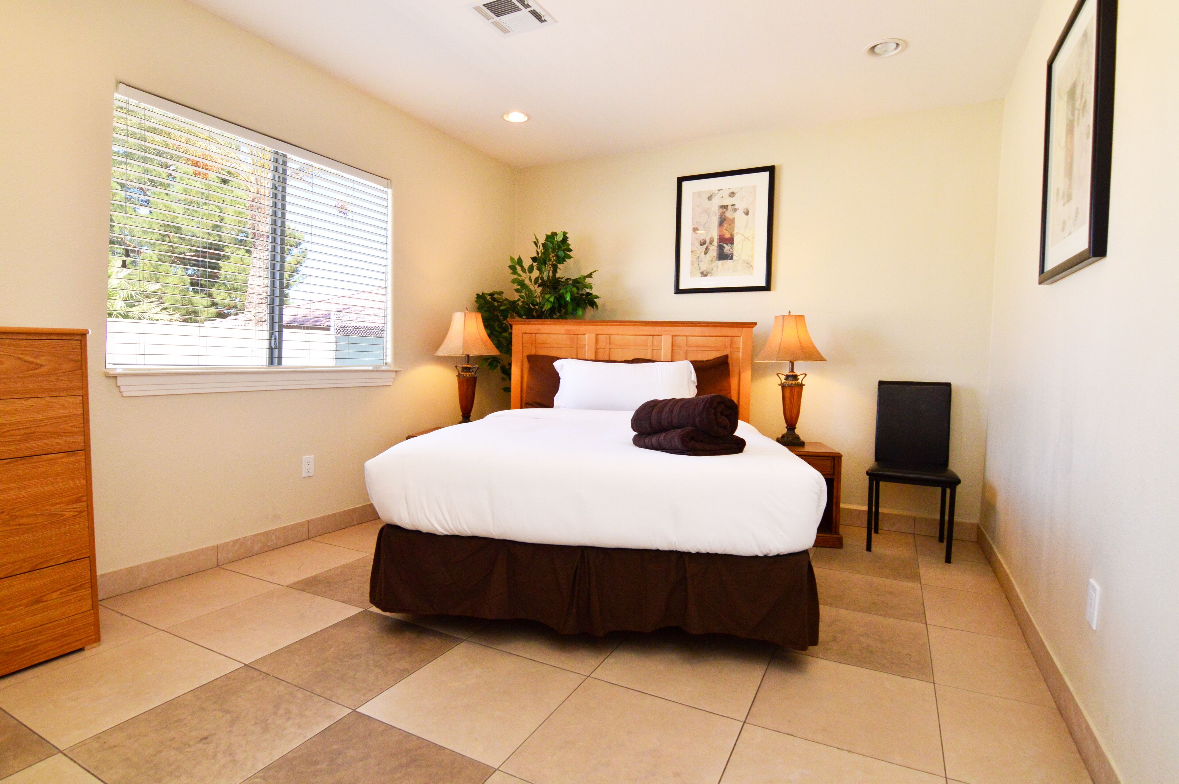 A hotel room with a large bed, two bedside lamps, a small table, a chair, and a window with blinds.
