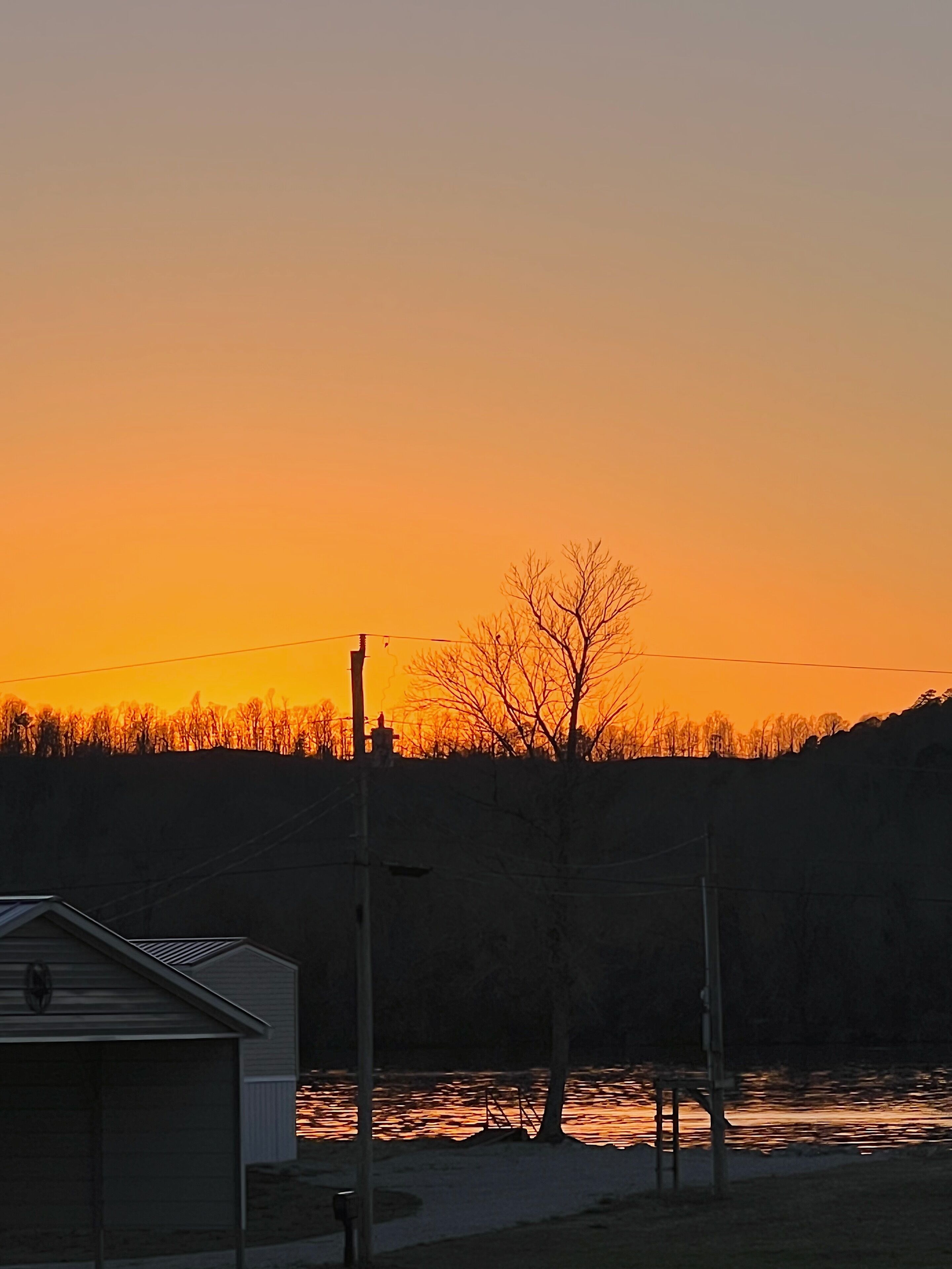 Un coucher de soleil sur une étendue d’eau, avec un bâtiment et des arbres sans feuilles au premier plan.