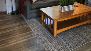 Een woonkamer met een grijze bank, een houten salontafel, een kamerbreed tapijt met een patroon, en een raam met zonwering.