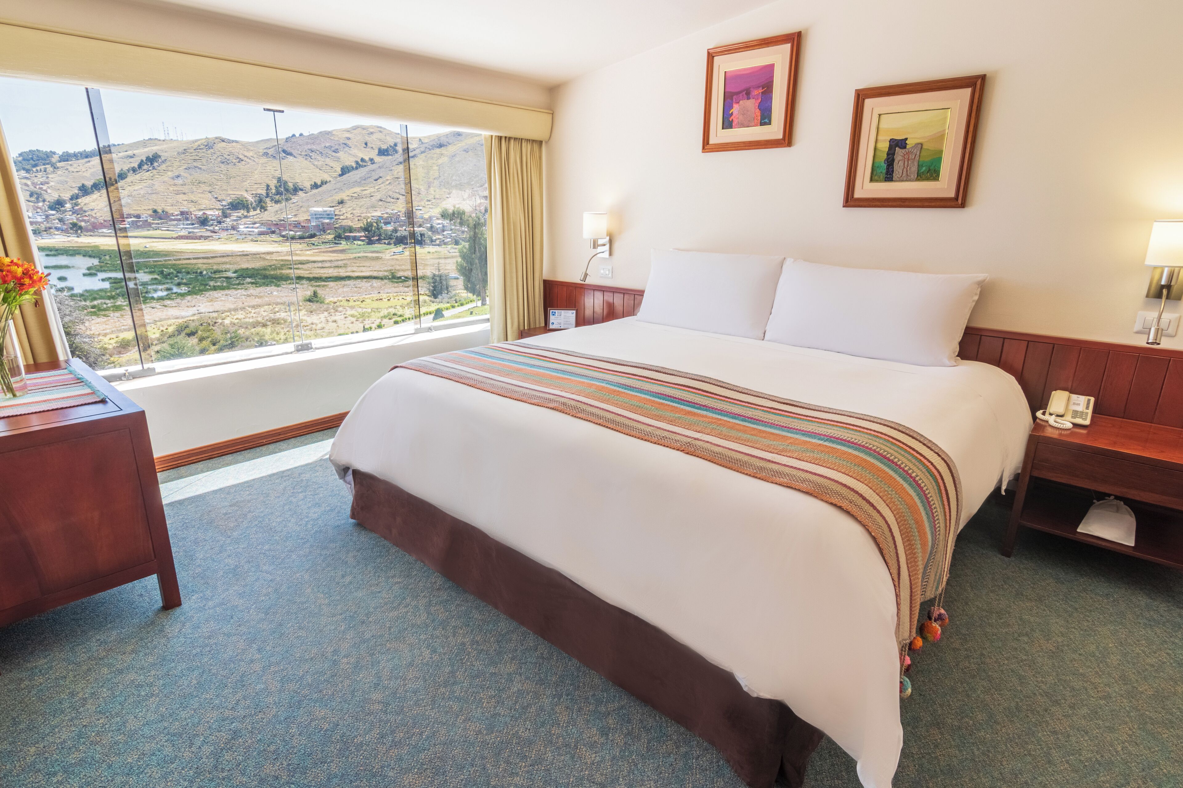 Ein Hotelzimmer mit einem großen Bett, einem Nachttisch, zwei gerahmten Bildern an der Wand und einem Fenster mit Blick auf ein Tal.