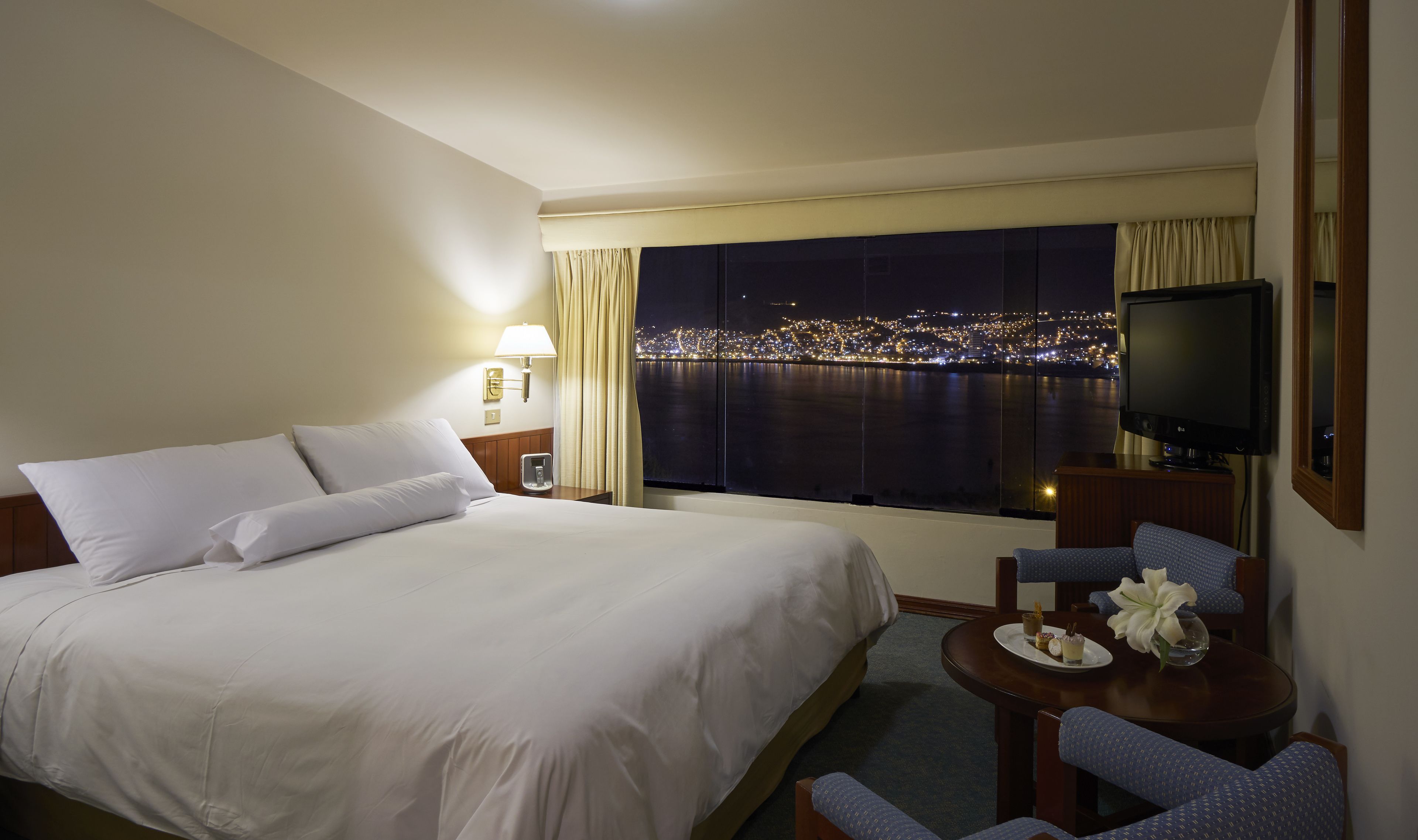 A hotel room with a large bed, a desk with a chair, a television, and a view of a cityscape at night.