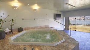 A modern bathroom with a large bathtub, a stone bench, a flat-screen TV mounted on the wall, and a view of the beach through large windows. - GHL Hotel Lago Titicaca (Puno)