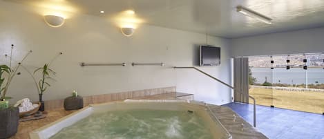 A modern bathroom with a large bathtub, a stone bench, a flat-screen TV mounted on the wall, and a view of the beach through large windows.