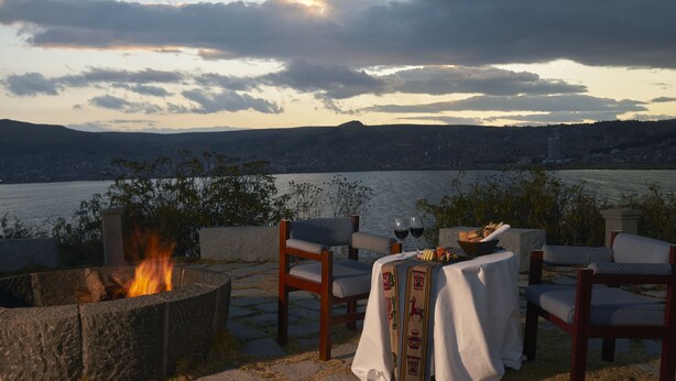 A fire pit with chairs and a table set for two, overlooking a body of water and a cityscape. - GHL Hotel Lago Titicaca (Puno)