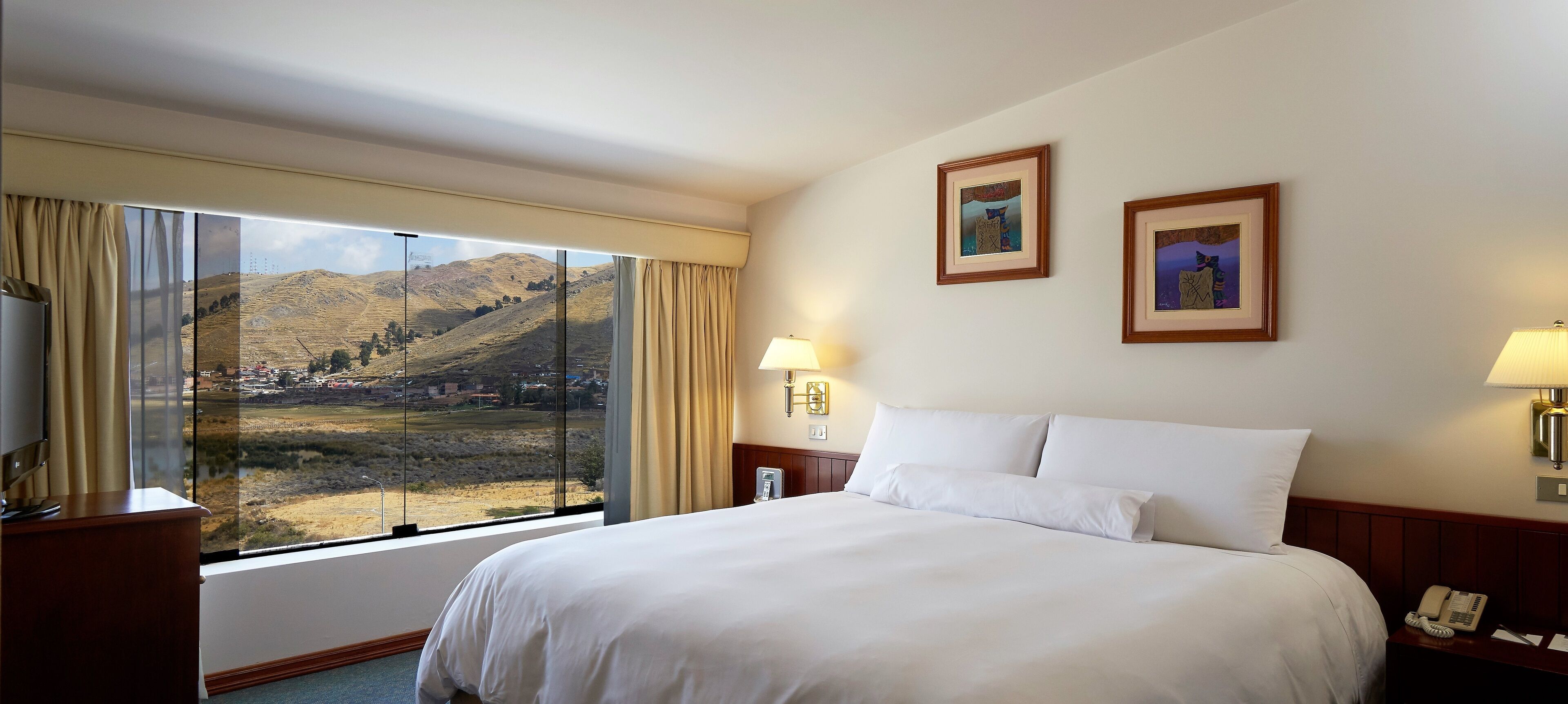Habitación de hotel con una cama grande, una mesita de noche con lámpara, un teléfono y dos cuadros en la pared.
