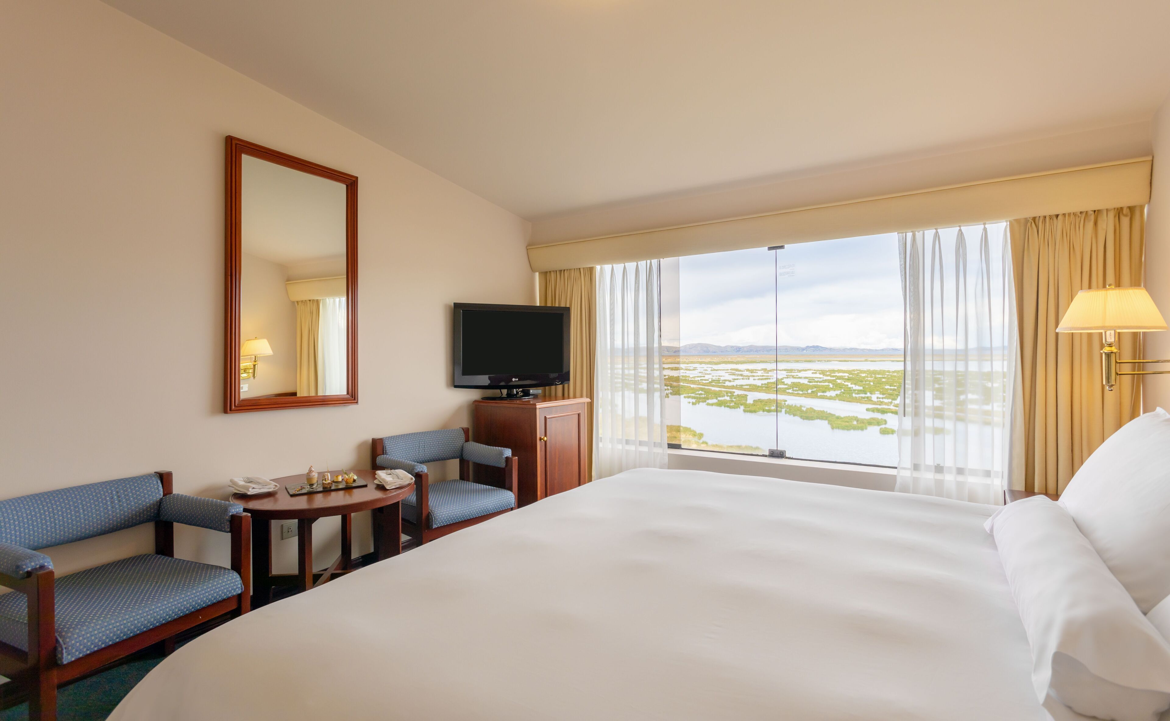 Habitación de hotel con una cama grande, un escritorio con una silla, televisión y vista a una masa de agua con montañas al fondo.