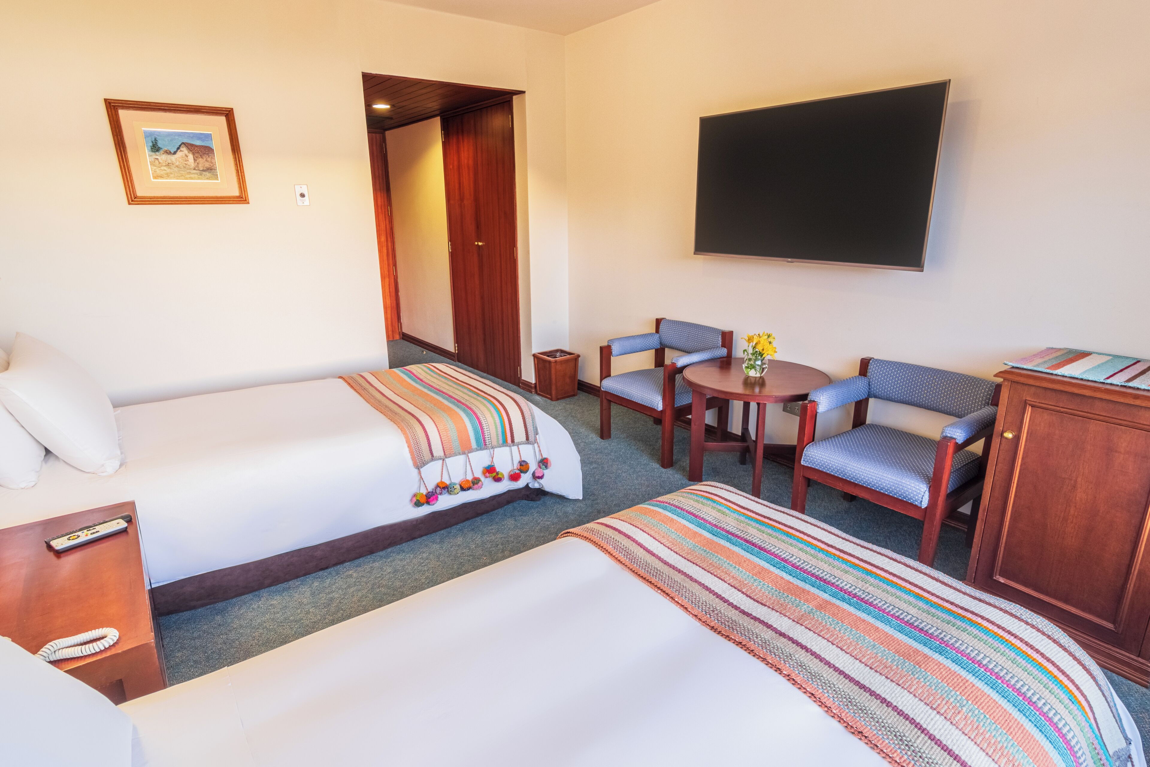 Ein Hotelzimmer mit zwei Betten, einem Fernseher, einem kleinen Tisch mit Stühlen und einer Tür zu einem weiteren Raum.