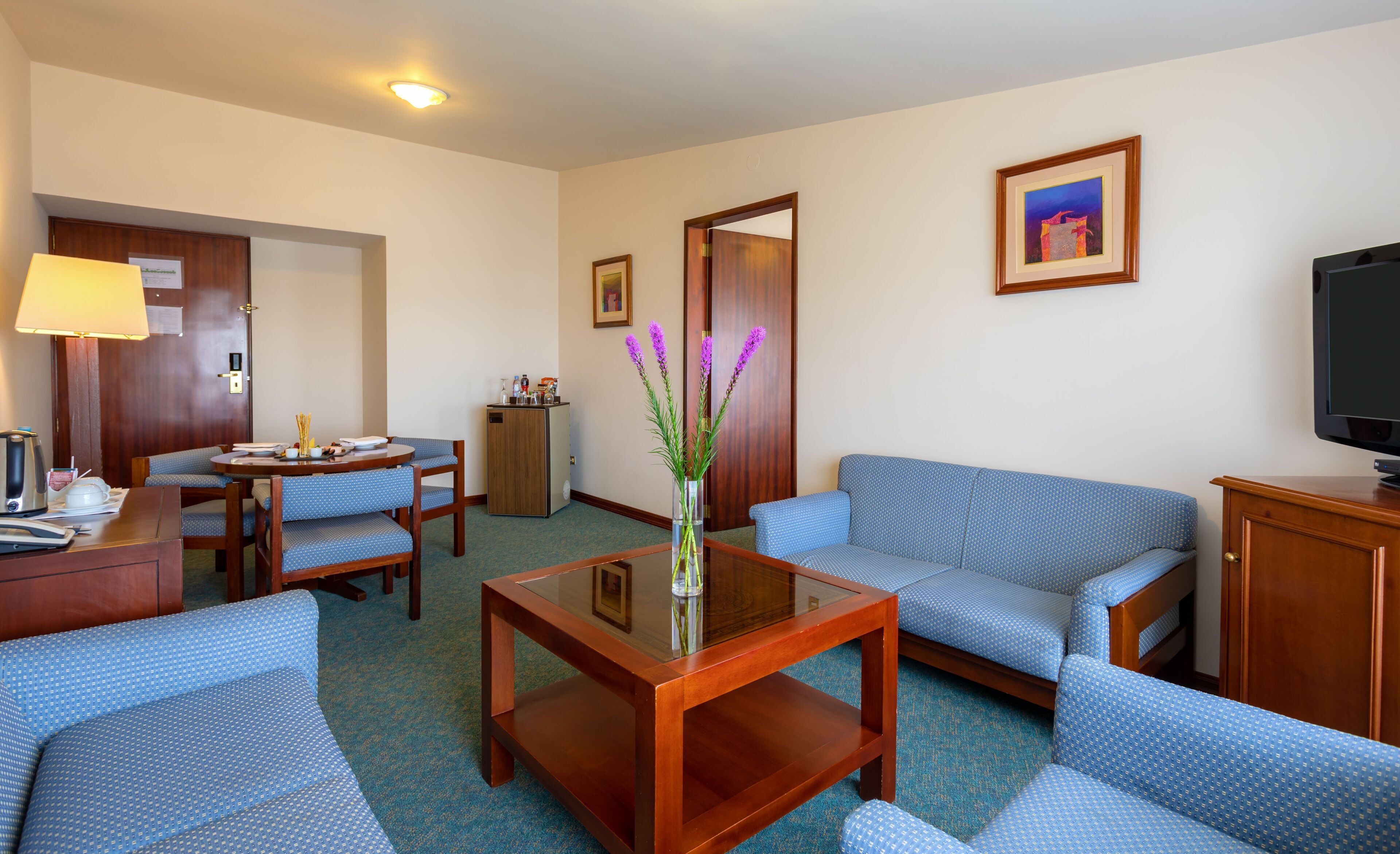 A hotel room with a sofa, chairs, a coffee table, a TV, a desk with a lamp, and a refrigerator.
