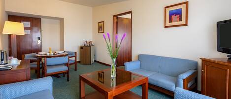 A hotel room with a sofa, chairs, a coffee table, a TV, a desk with a lamp, and a refrigerator.