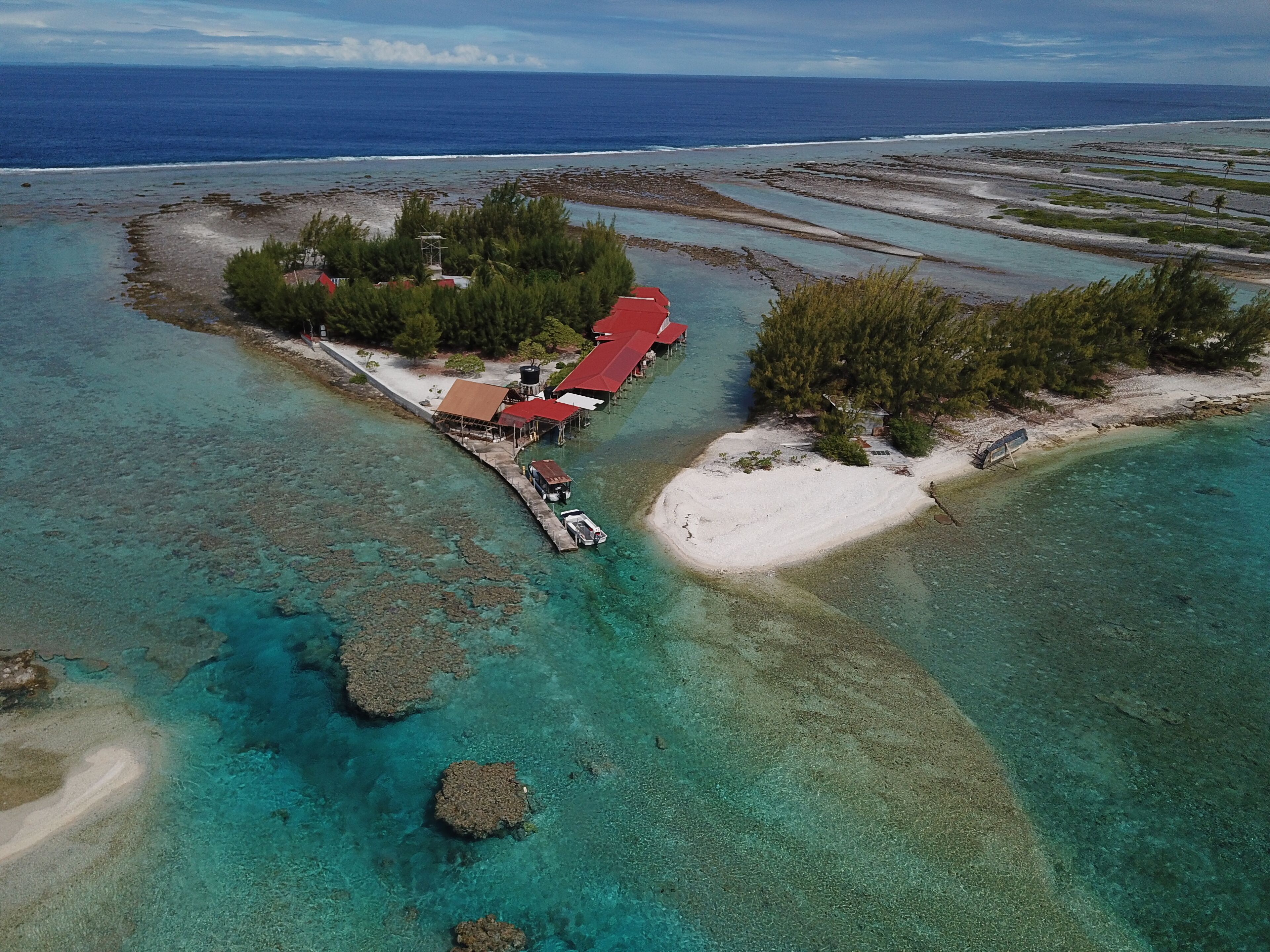 An aerial view of a coastal property with a pier leading to a building with a red roof, surrounded by clear turquoise waters and a sandy beach.