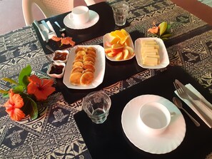 A table set with a white cup, a glass of water, a plate of fruit, a tray of pastries, and a small bowl of jam. - Pension Motu Aito Paradise (Fakarava)