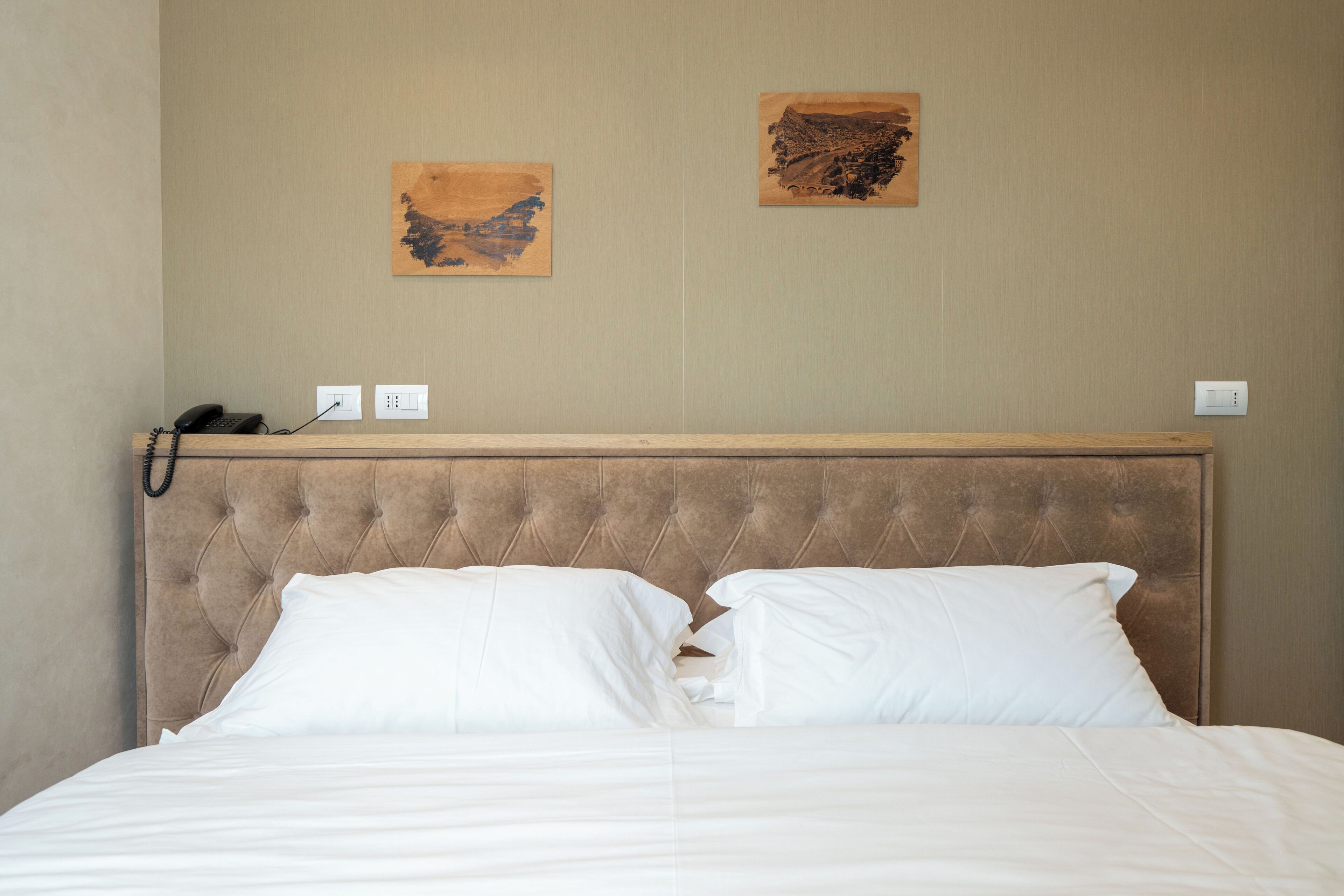 A neatly made bed with a tufted headboard, two wall-mounted artworks, and a telephone on the nightstand.