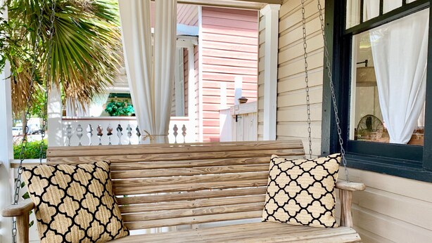 A wooden porch swing with two patterned cushions, a white railing, and a view of a pink building in the background. - The RED DOOR: Explore Pensacola's Downtown Entertainment next to Wedding Venues! (Pensacola)