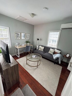Un salón moderno con un sofá, una mesa de centro y un escritorio con una silla. Hay un televisor sobre un mueble bajo y obras de arte en las paredes.