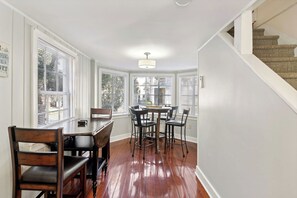 Comedor con mesa y sillas de madera, una lámpara de techo y ventanas grandes con persianas.