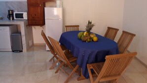 A small dining area with a table covered by a blue tablecloth, surrounded by wooden chairs. A refrigerator and a microwave are visible in the background.