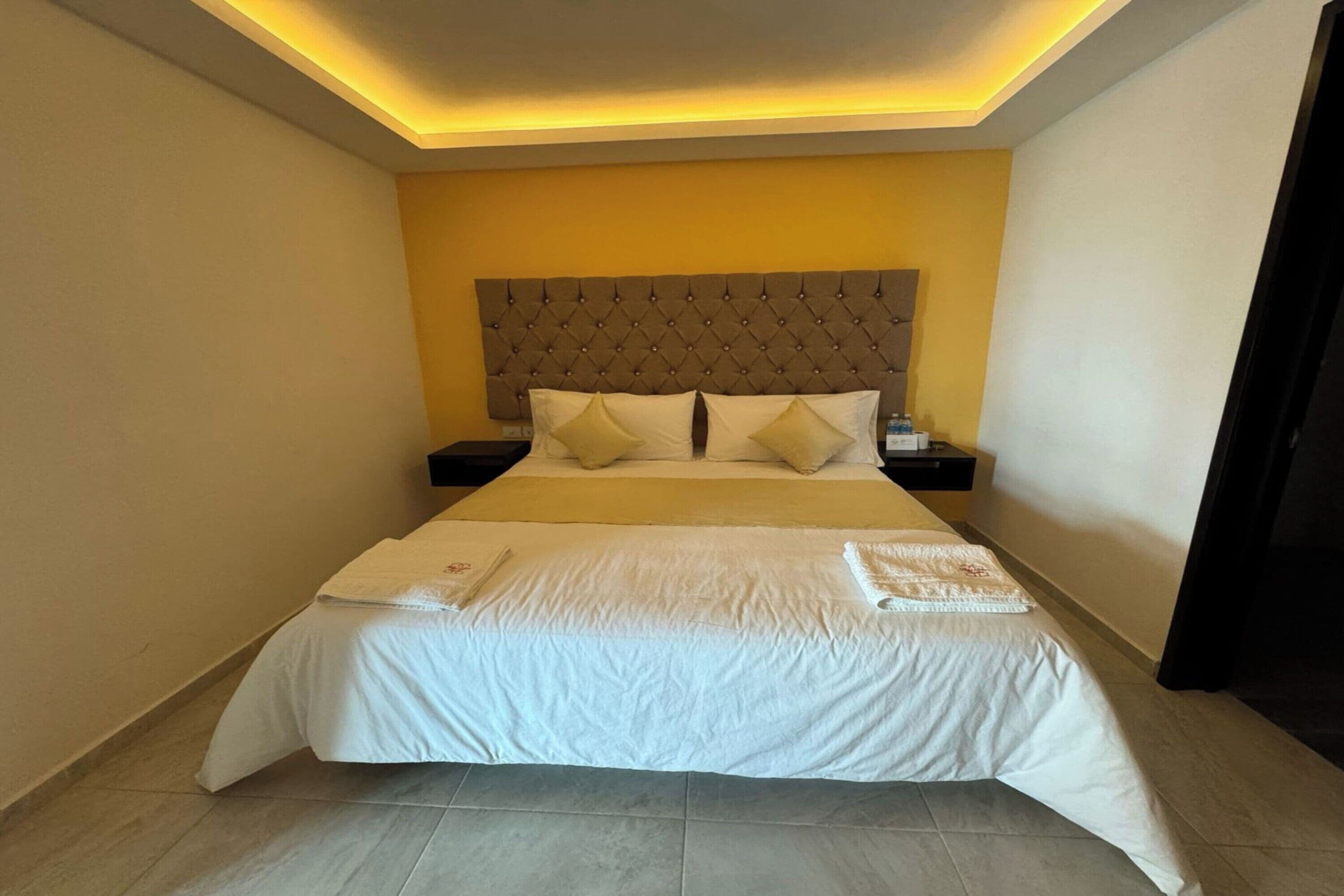Una cama bien tendida con un cabecero acolchado, ropa de cama blanca y una mesita de noche con una lámpara.