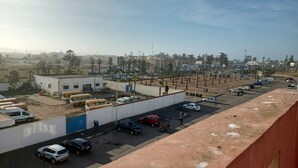 Un estacionamiento con varios coches y autobuses, un edificio con puerta azul y un cielo despejado.