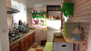 Cocina compacta con gabinetes de madera, estufa a gas y refrigerador. Se observan repisas con utensilios de cocina y una pequeña mesa con mantel de lunares.