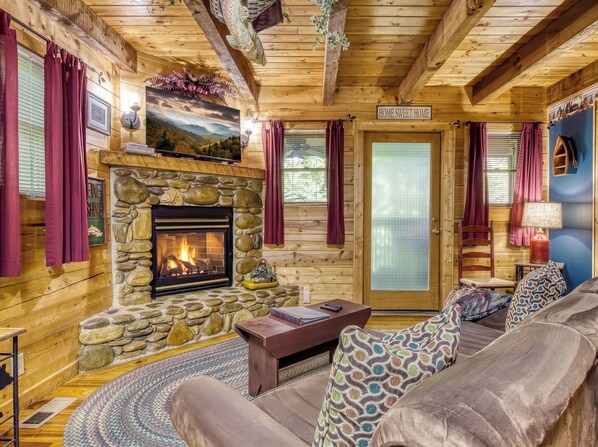 L’intérieur d’un chalet accueillant, avec une cheminée en pierre, des murs en bois, un canapé et une porte sur laquelle on peut lire : « Chez soi, on est bien ».