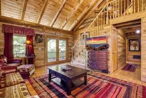 Interior de una cabaña de troncos con una escalera, chimenea y un ventanal grande.