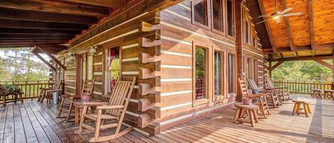 A log cabin with a deck furnished with chairs and tables, large windows, and a wooden ceiling.