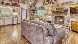 A log cabin interior with a fireplace, leather sofa, kitchen area, and wooden ceiling.