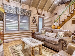 L’intérieur d’une cabane en rondins, avec un canapé en cuir, une table basse en bois, un escalier et une fenêtre équipée de stores.