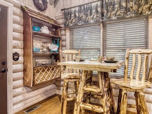 L’intérieur d’un chalet rustique, avec une table et des chaises en bois, une étagère ornée d’objets décoratifs et une fenêtre avec des stores.