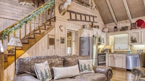 A cozy cabin interior with a leather sofa, wooden staircase, kitchen area, and a mounted deer head.