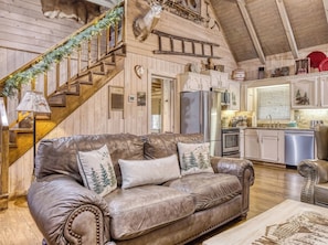 A cozy cabin interior with a leather sofa, wooden staircase, kitchen area, and a mounted deer head.