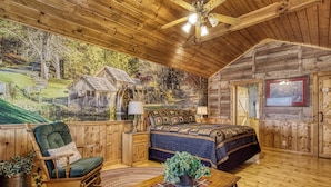 Interior de una cabaña de troncos con cama, mecedora y un mural de paisaje rural.