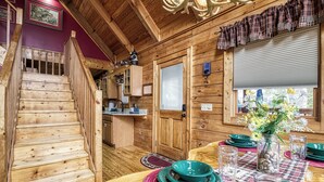 Interior de una cabaña de madera con escalera, un comedor con mesa dispuesta para dos personas y una zona de cocina.