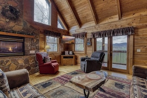 A log cabin interior with a stone fireplace, wooden walls, a television, and a view of mountains through large windows.