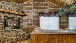 Interior de una cabaña de troncos con chimenea de piedra, una bañera de madera y una ventana con persianas.