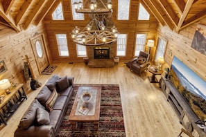 A spacious log cabin interior with a large wooden ceiling, a central fireplace, a sofa, a coffee table, and a painting on the wall.