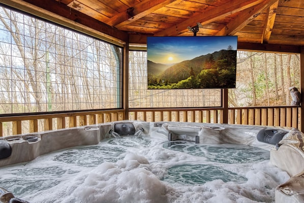 A hot tub area with wooden beams, a large window, and a television displaying a mountain landscape.