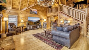 A log cabin interior with a wooden staircase, a fireplace, a large leather sofa, and a mounted animal head.