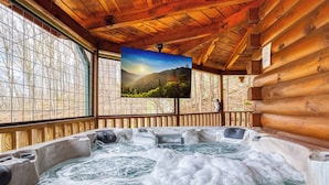 A wooden cabin with a hot tub, a large window, and a television displaying a mountain scene.