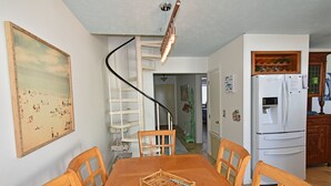 Un coin repas avec une table et des chaises en bois, un escalier en colimaçon et une cuisine équipée d’un réfrigérateur.
