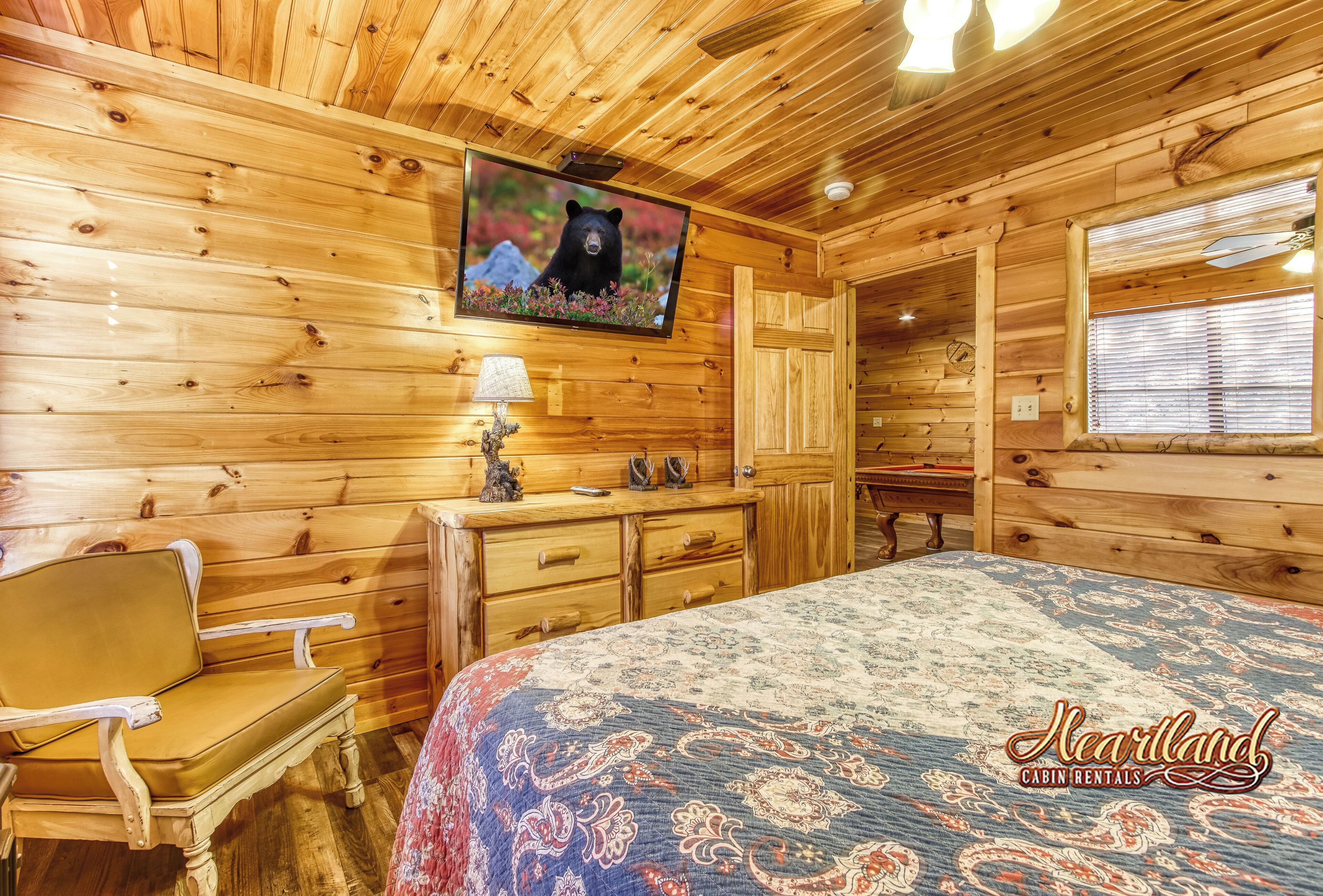 A cabin with a wooden interior, a bed with a patterned bedspread, a television mounted on the wall, a wooden dresser, and a bench.