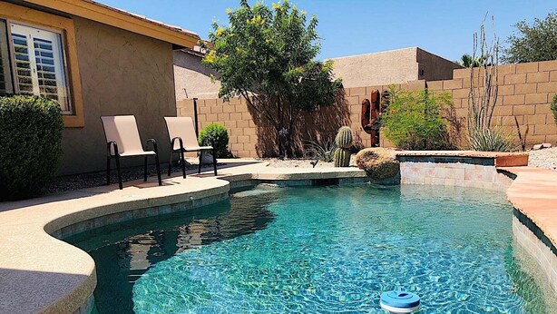 Una piscina cristallina con un dispositivo galleggiante blu, circondata da un muro in mattoni e un piccolo giardino con un albero e degli arbusti.