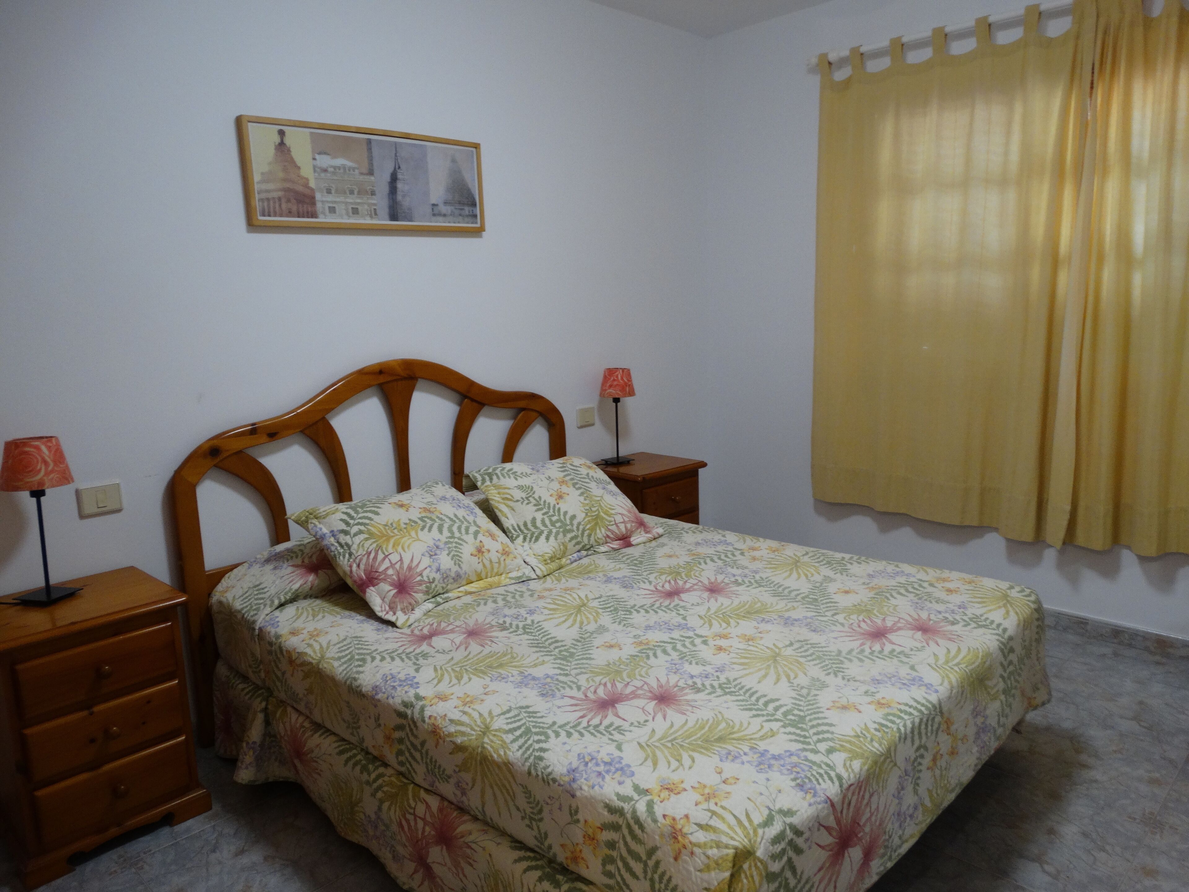 Un dormitorio con cama de madera, ropa de cama floral, dos mesitas de noche con lámparas, un cuadro en la pared y una cortina amarilla.