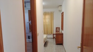 Un couloir étroit avec une porte en bois sur la droite, un lit surmonté d’un rideau jaune et une commode en bois.
