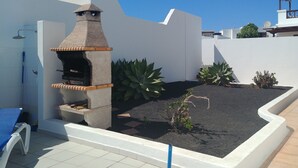 Une terrasse avec un barbecue en pierre, une chaise bleue et un petit espace jardin agrémenté de plantes et de gravier.