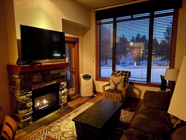 A living room with a stone fireplace, a flat-screen TV, a comfortable couch, and a view through large windows.