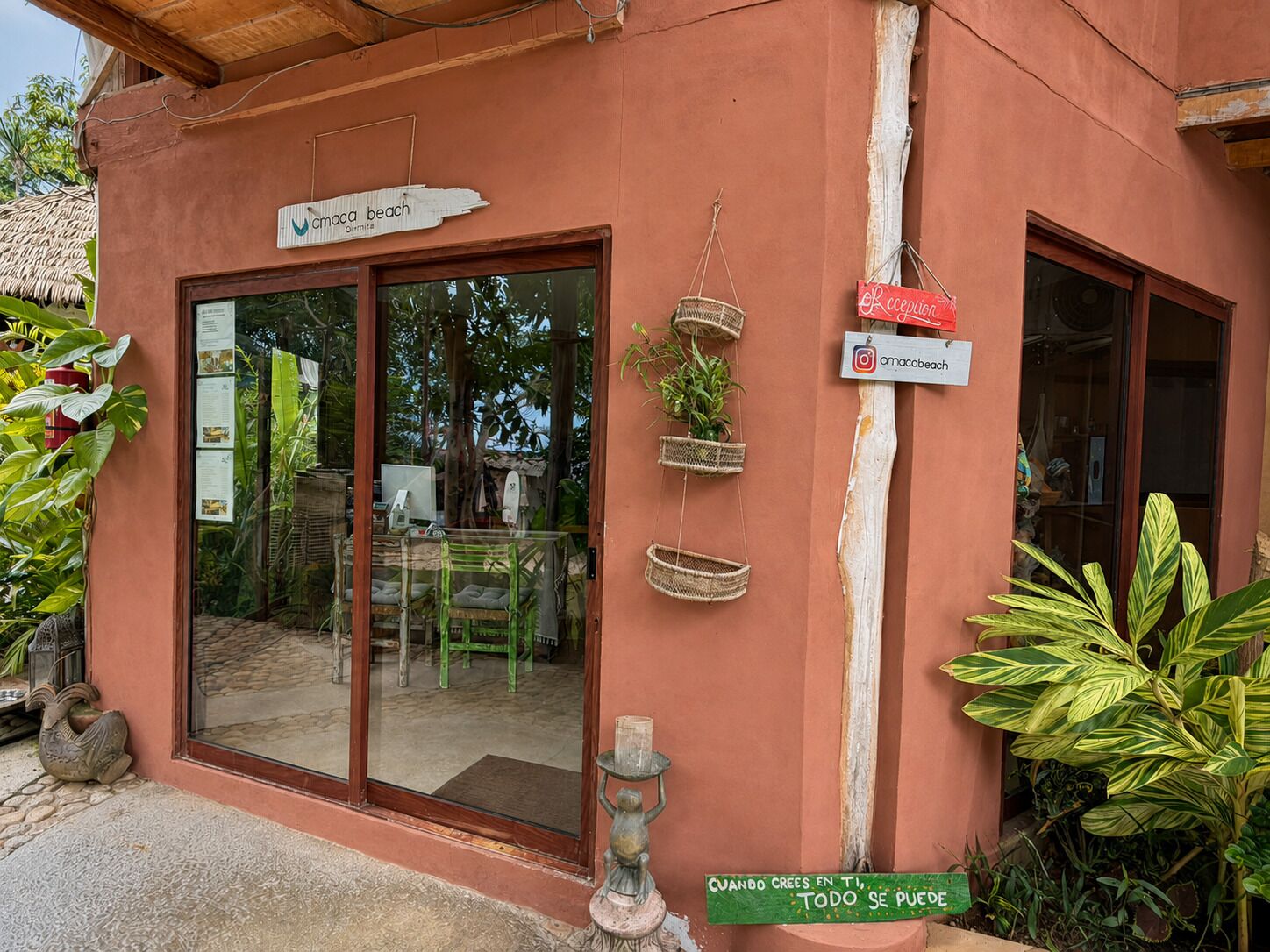 Un edificio con entrada por puerta de cristal, un cartel con el nombre 'Cmooca beach' y un pequeño espacio exterior con un banco y plantas en macetas.