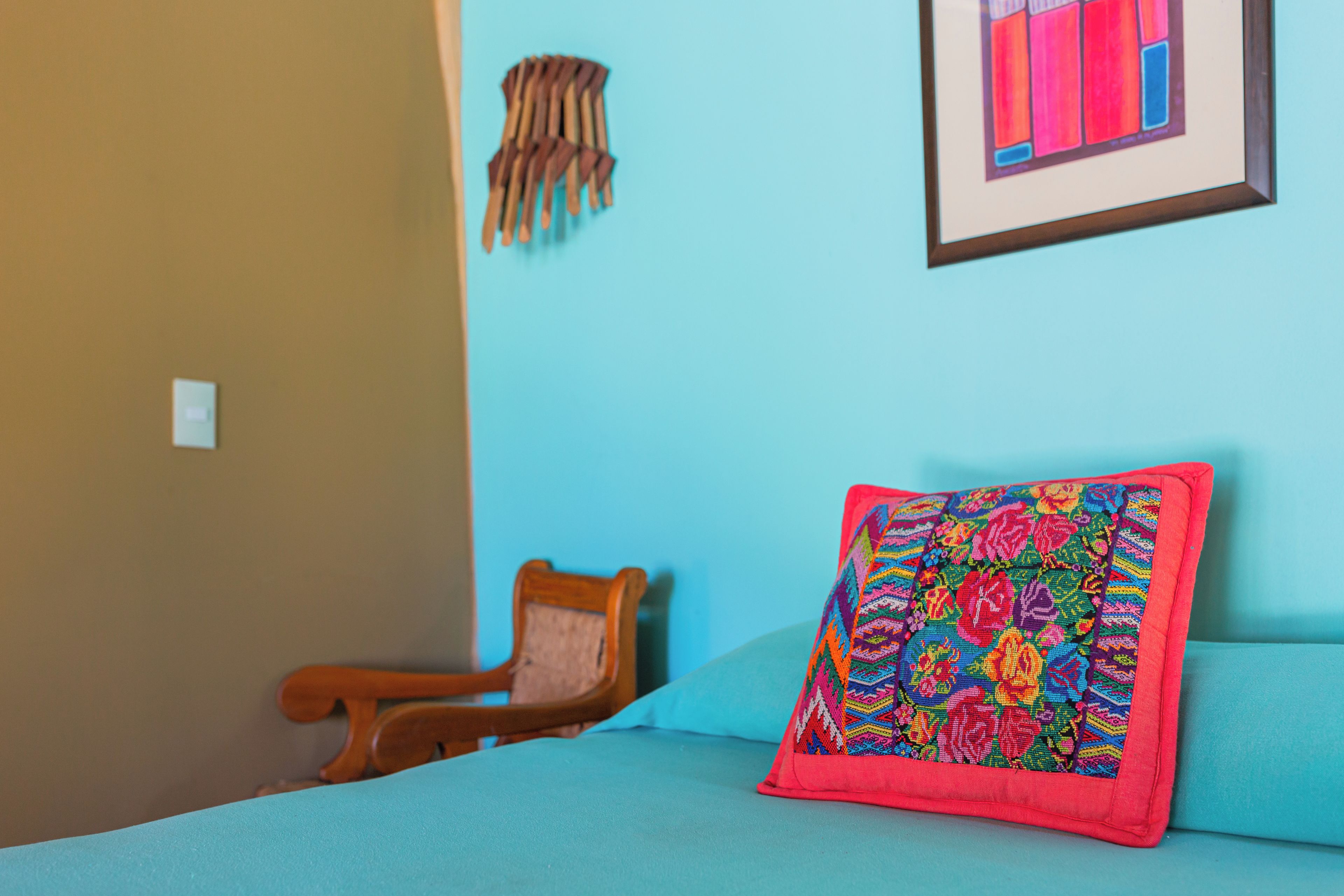 A room with a bed, a chair, a wall with a painting, and a wall-mounted item.