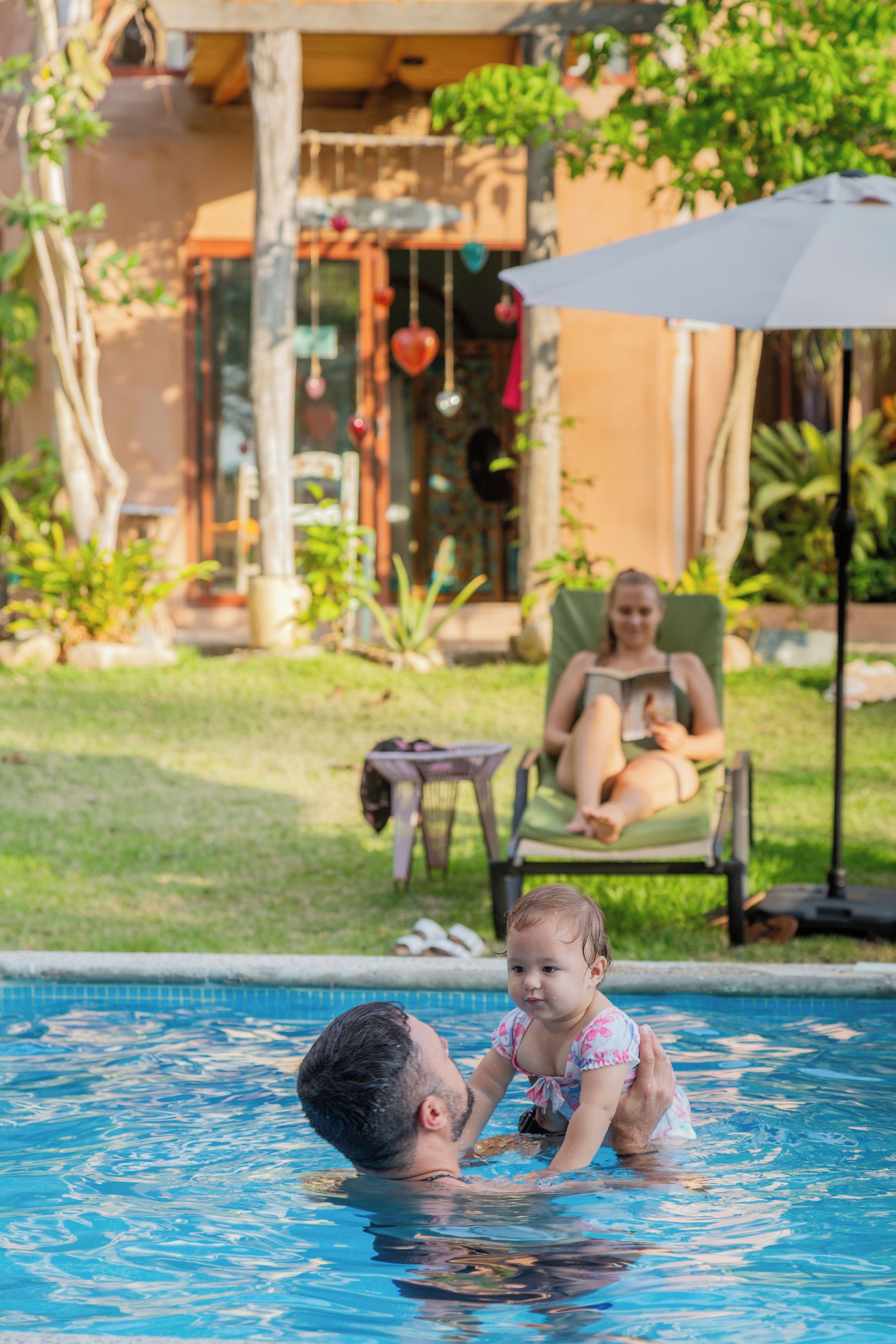 Una piscina con una persona y un niño en el agua, una silla de descanso y una zona cubierta con plantas.